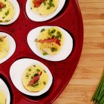 Deviled Eggs on a red dimpled platter on bamboo cutting board with chives and bacon