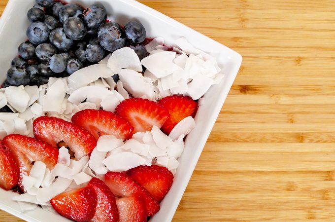 Red white blue acai bowl. blueberries, sliced strawberries, coconut flakes in white bowl on bamboo cutting board