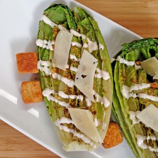 Grilled Caesar Salad drizzled with dressing, croutons, shaved parmesan cheese on white plate on bamboo cutting board