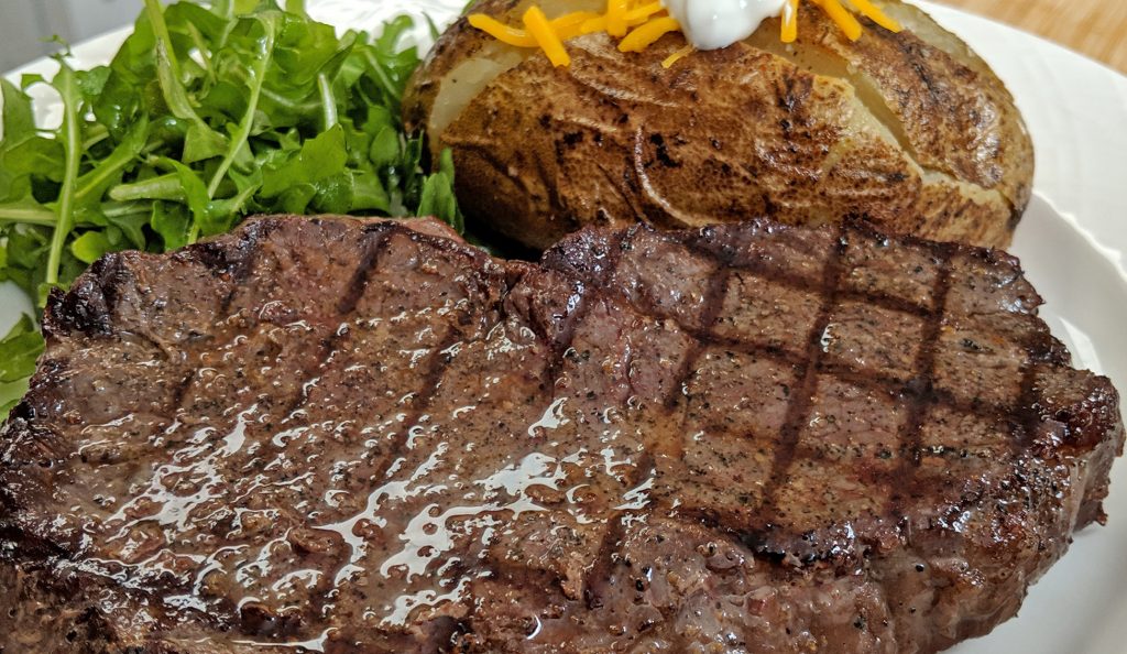 A steak with diamond sear marks on a white plate with a baked potato and arugula salad