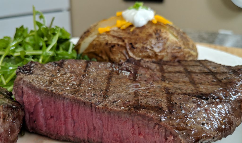 Cut steak cooked medium to 145 degrees on a white plate iwth a baked potato, sour cream, cheddar cheese, chives and an arugula salad.