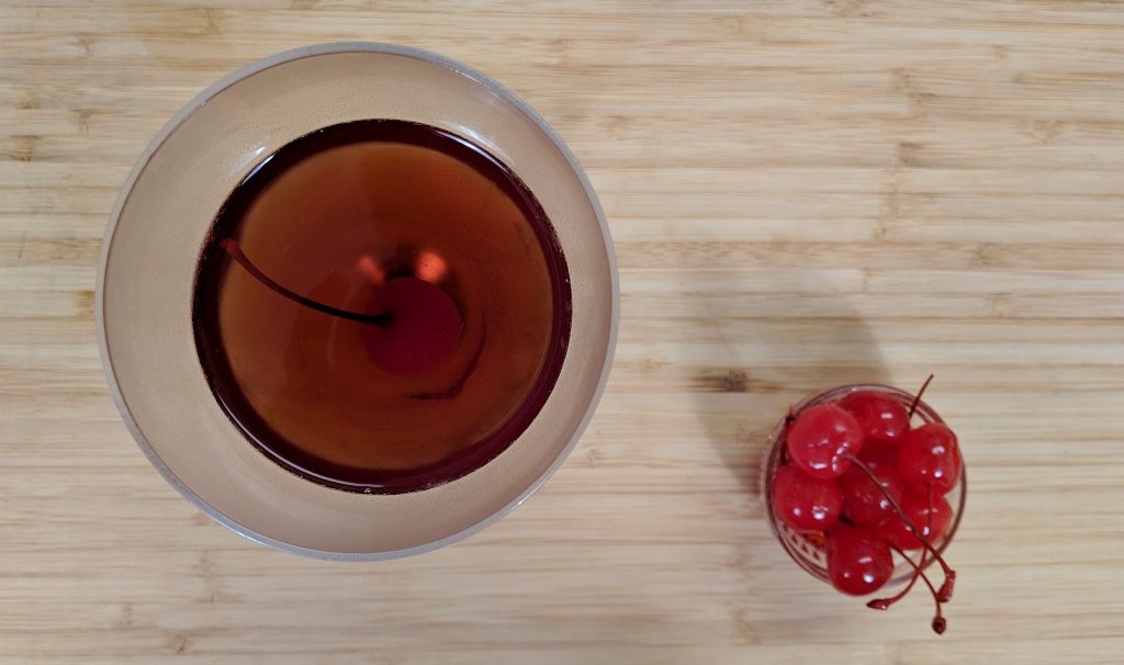 Ovehead view of manhattan cocktail in nick and nora glass with maraschino cherries on a bamboo cutting board